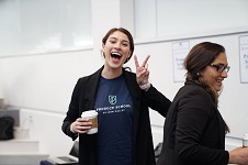 A picture of two women in dark clothes in an office, one of them showing the V sign and holding a paper cup of coffee.